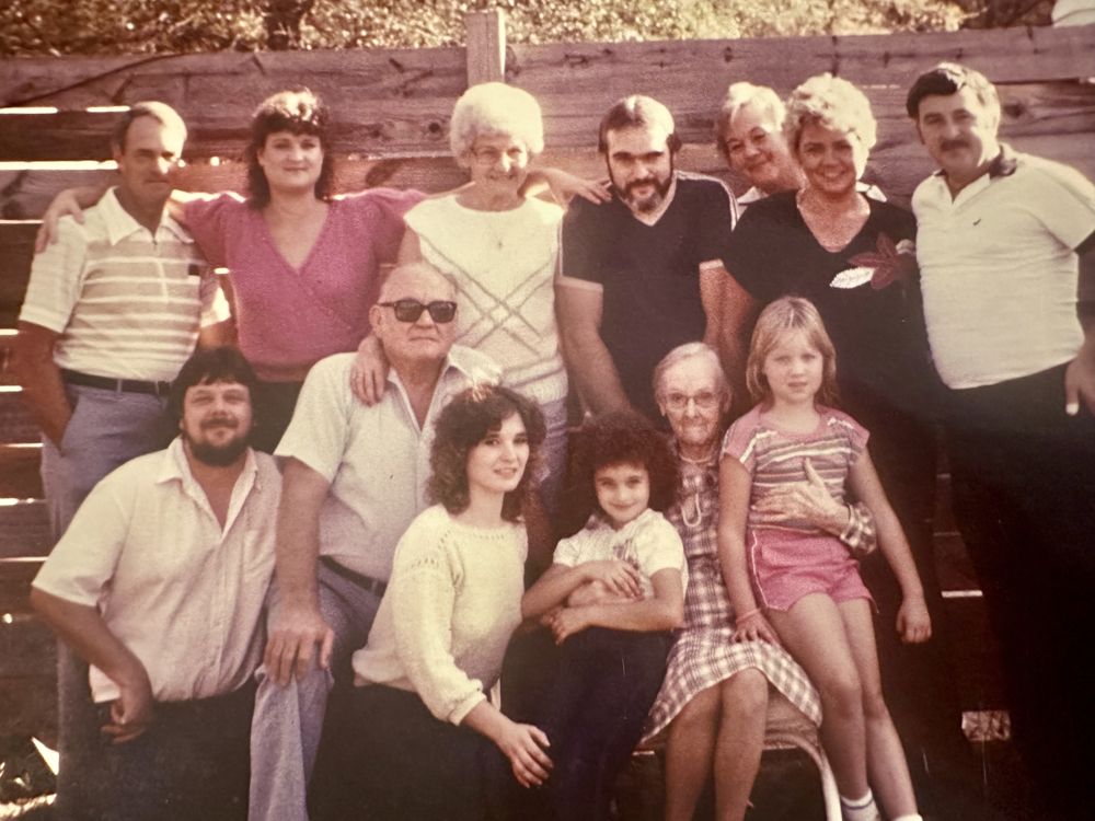 Family group photo outdoors in front of a wooden fence. Bobby stands at top left, his daughter Cheryl beside him, and his son Shane at top center.